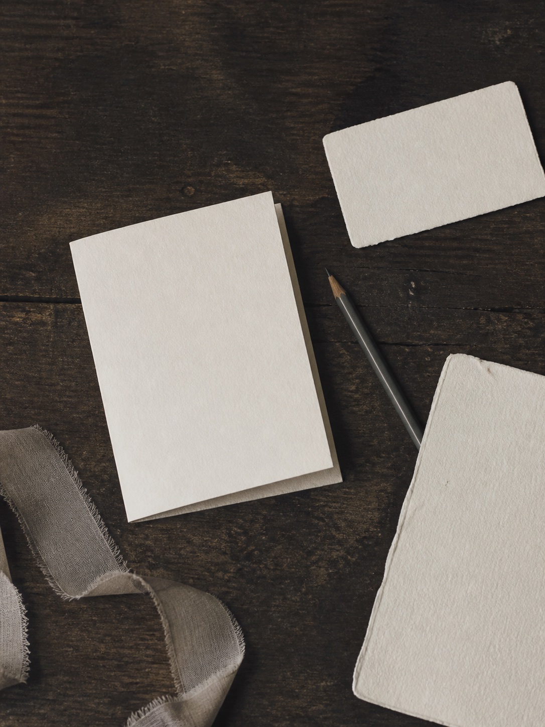 Blank ivory paper pieces, a graphite pencil and frayed linen ribbon arranged on a dark wooden table.