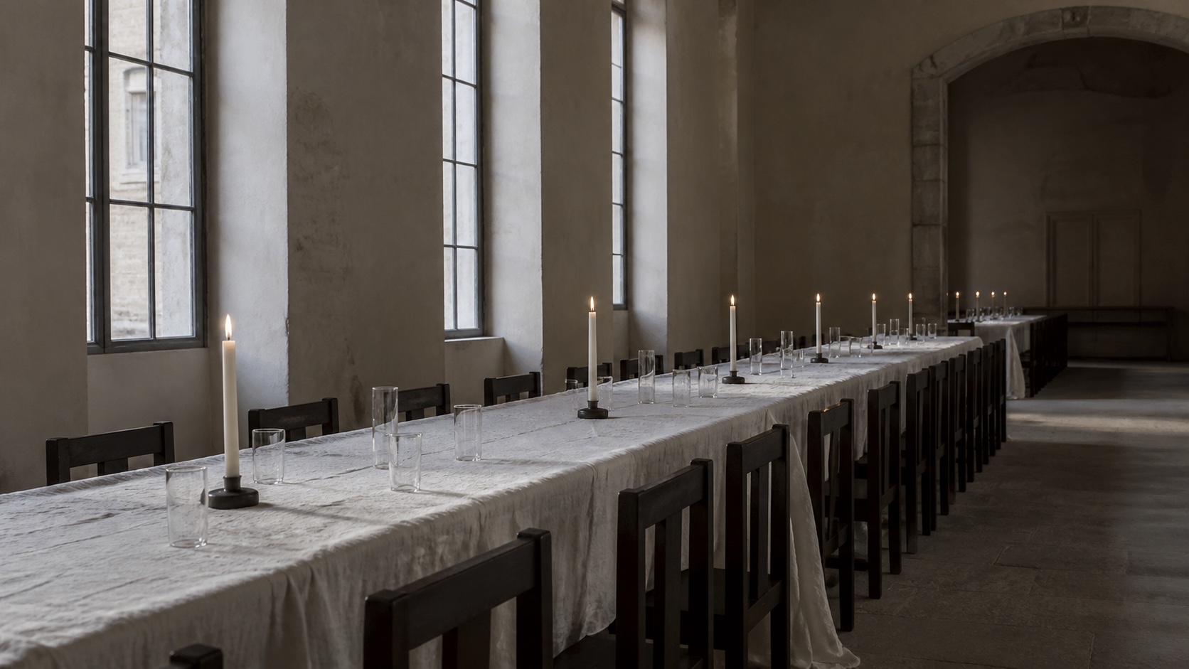 A long empty table covered in white linen, with dark straight-backed chairs, clear glasses and tall white candles in a stone hall.