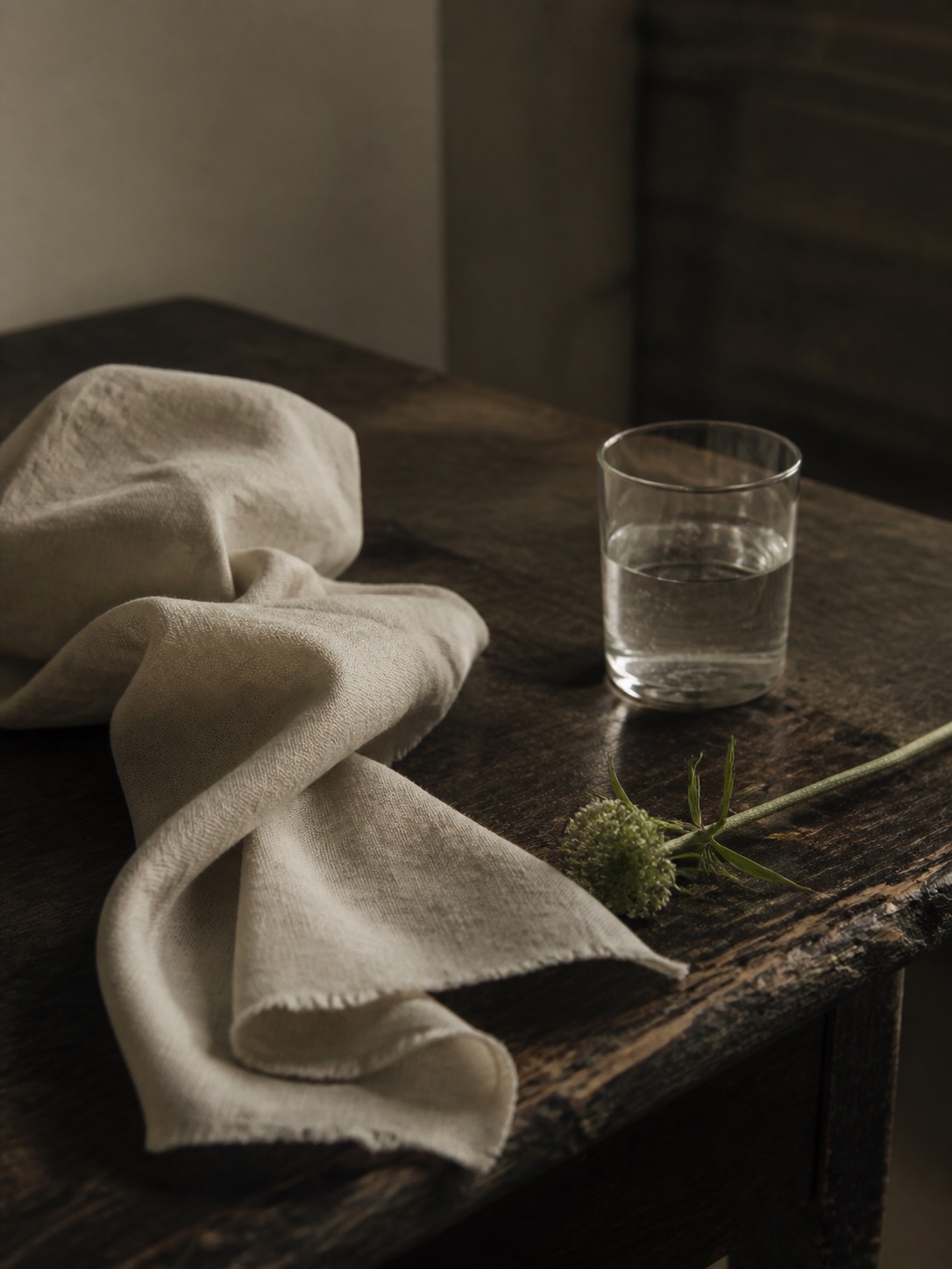 Rumpled ivory linen, a water glass and a clipped green stem on a worn dark table.